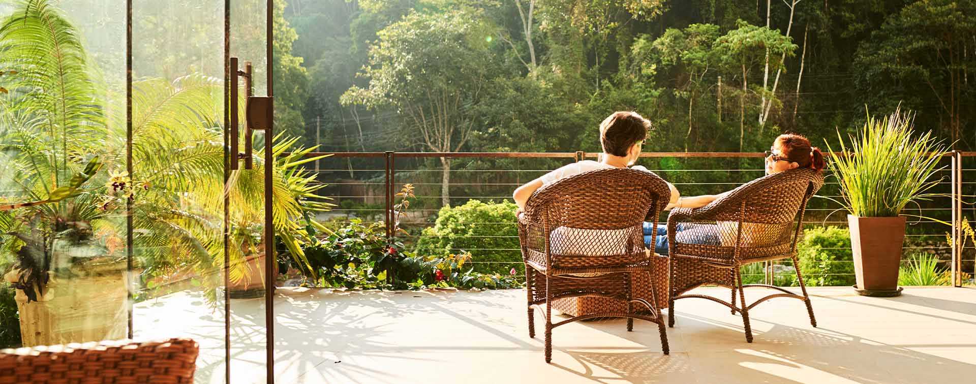 A couple relaxing on a sunlit terrace surrounded by lush greenery, overlooking a serene forest view. The scene evokes a sense of luxury, climate resilience, and investment in tranquil, nature-rich locations.