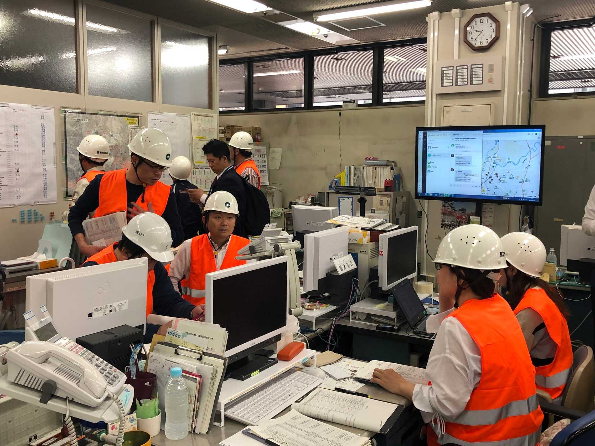 Workers in a control room wearing helmets and safety vests, monitoring screens and maps during an emergency or operational task.